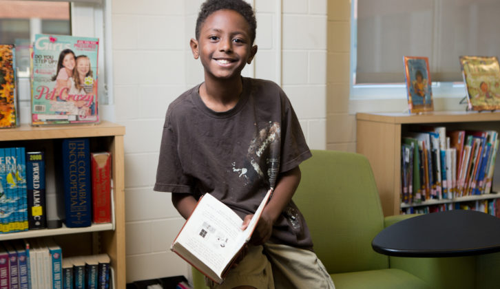 黑人男性中学生，带着书坐在椅子扶手上，微笑着阅读识字计划的文章