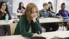 在一间学生教室里，穿着绿色毛衣的女孩低头看着桌子，寻找如何解决学生压力和行为的文章