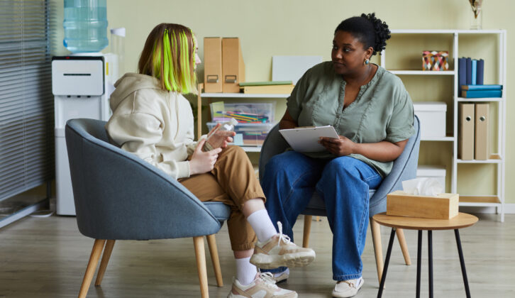 黑人女性的社会工作者和十几岁的女孩当他们坐在扶手椅在心理健康支持办公室的文章