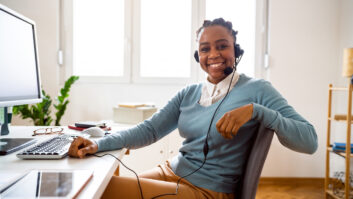 一名身穿淡蓝色毛衣、戴着有线耳机的黑人妇女坐在桌子上的电脑前，微笑着看一篇关于教育工作者微证书的文章。