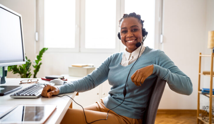 黑人妇女在淡蓝色的毛衣穿有线耳机在办公桌和电脑前微笑为教育者microcredentials文章。
