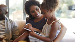 黑人女儿坐在黑人母亲的腿上。孩子有平板电脑,妈妈是指向东西。对于文章成功的读写程序